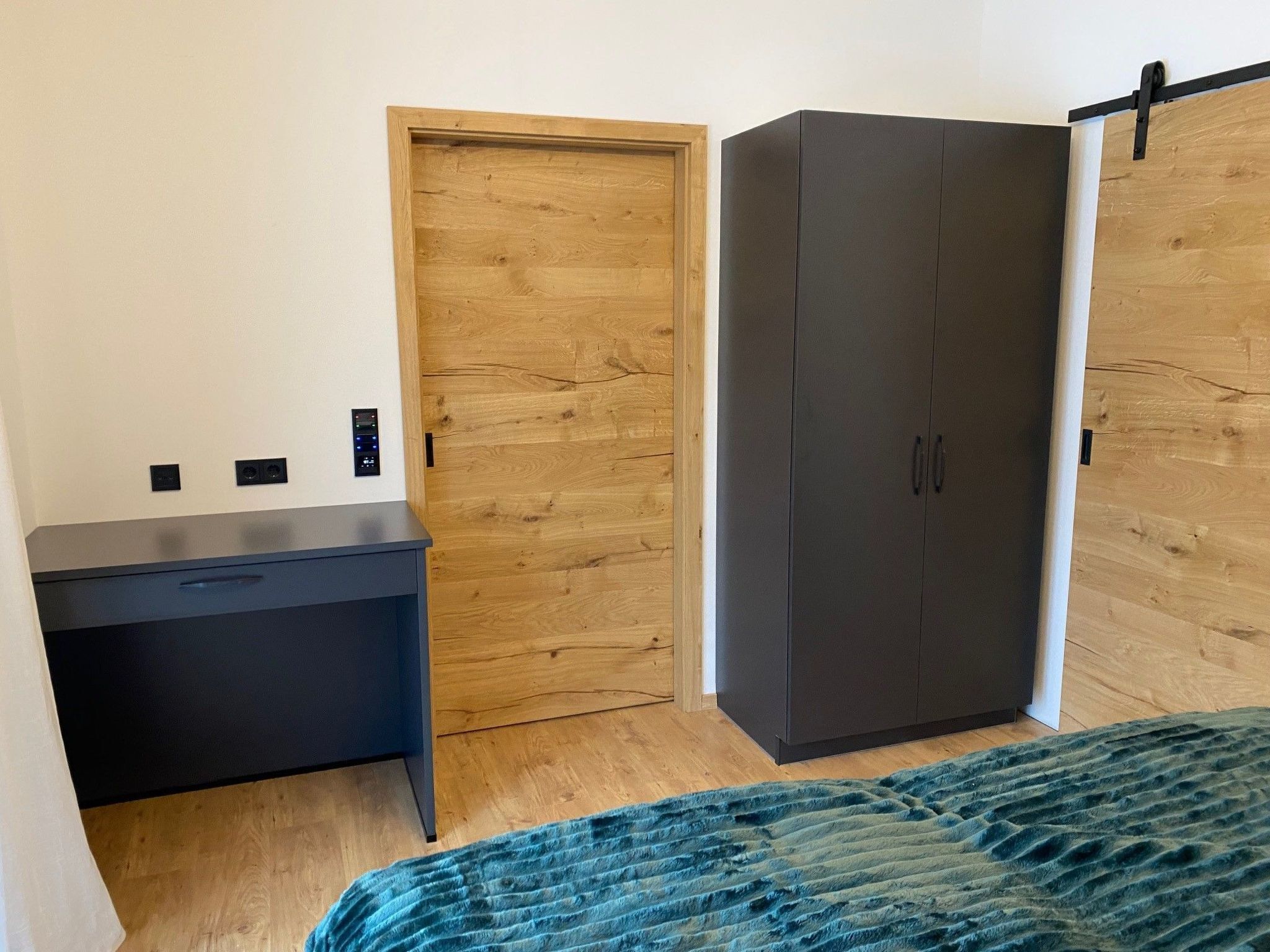 A bedroom with a wooden door, a dark-colored wardrobe, a desk, and a bed with a blue patterned bedspread.