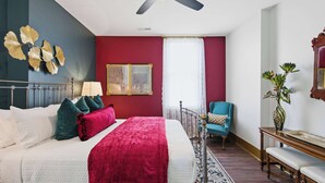 Een slaapkamer met een bed, een plafondventilator, een raam met gordijnen, een stoel en een nachtkastje met een vaas.