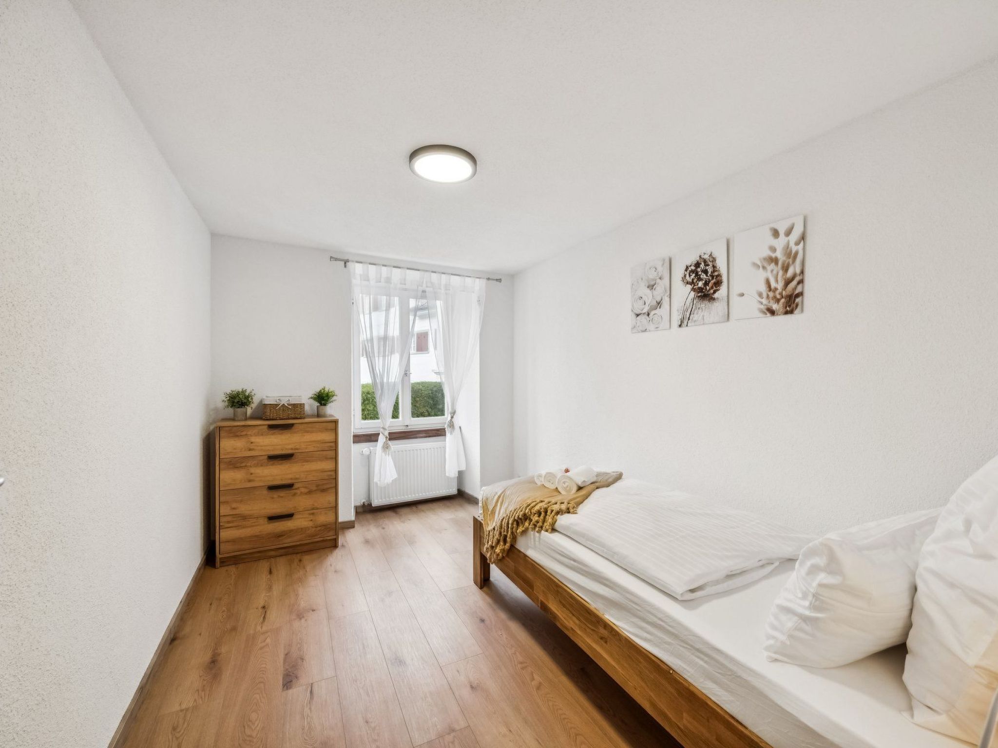 Een slaapkamer met een houten dressoir, een bed met witte beddengoed, een radiator en een raam met dunne gordijnen.