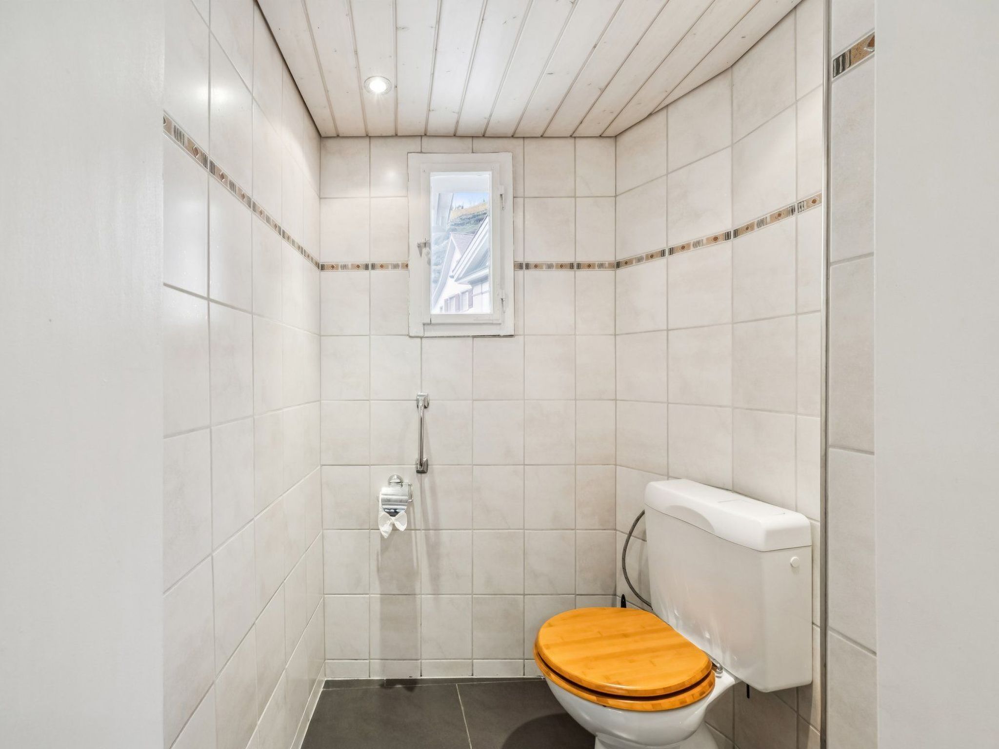 Ein kleines, kompaktes Badezimmer mit weißen Fliesen, einem hölzernen Toilettendeckel und einem Fenster über der Toilette.