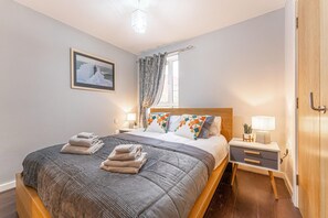 Un lit bien fait, avec une couette grise et deux oreillers, une table de chevet avec une lampe, et un tableau encadré au mur.