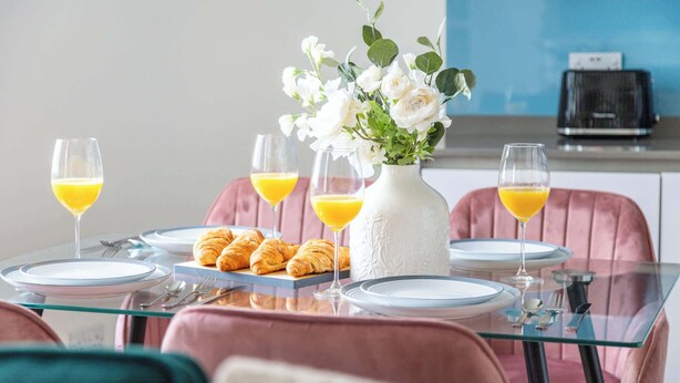 Een ontbijtsetting met een glazen tafel, roze stoelen, een vaas met bloemen, croissants en sinaasappelsap.