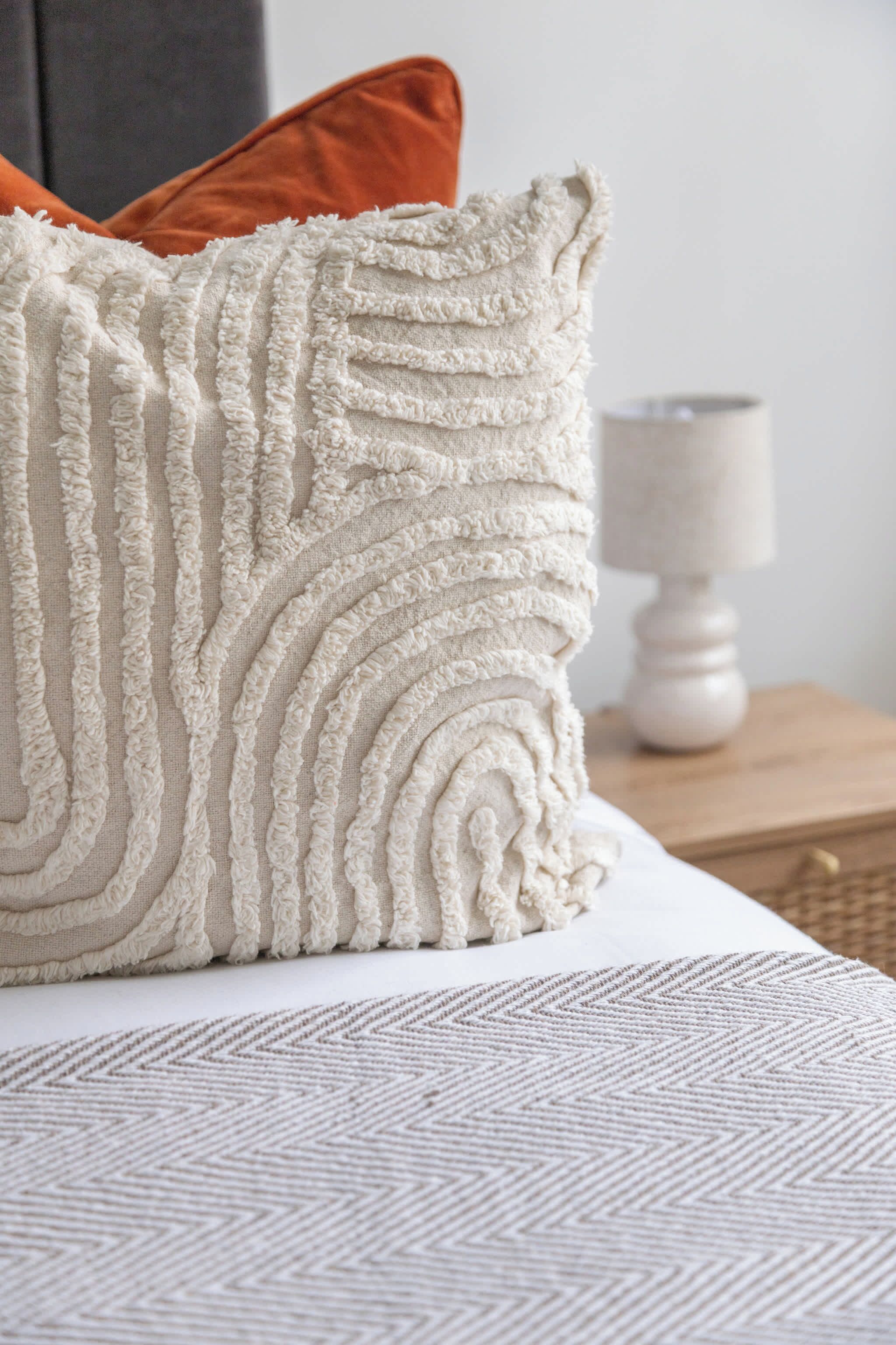A bed with a textured white pillow, a geometric patterned blanket, and a wooden nightstand with a white lamp.