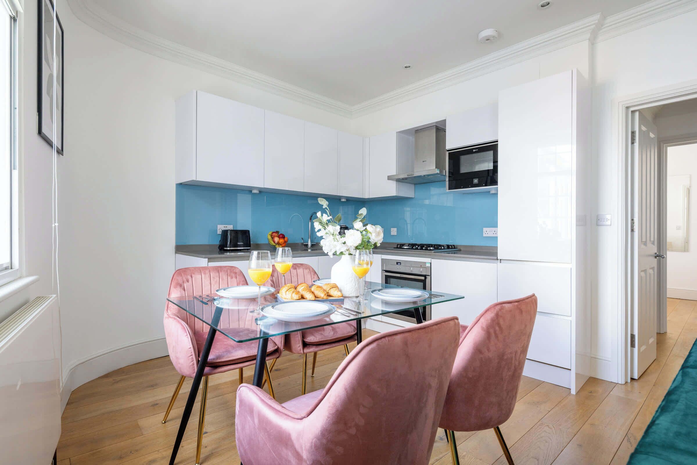 Une cuisine moderne avec des meubles blancs, un pare-feu bleu et un coin repas avec des chaises roses et une table en verre.