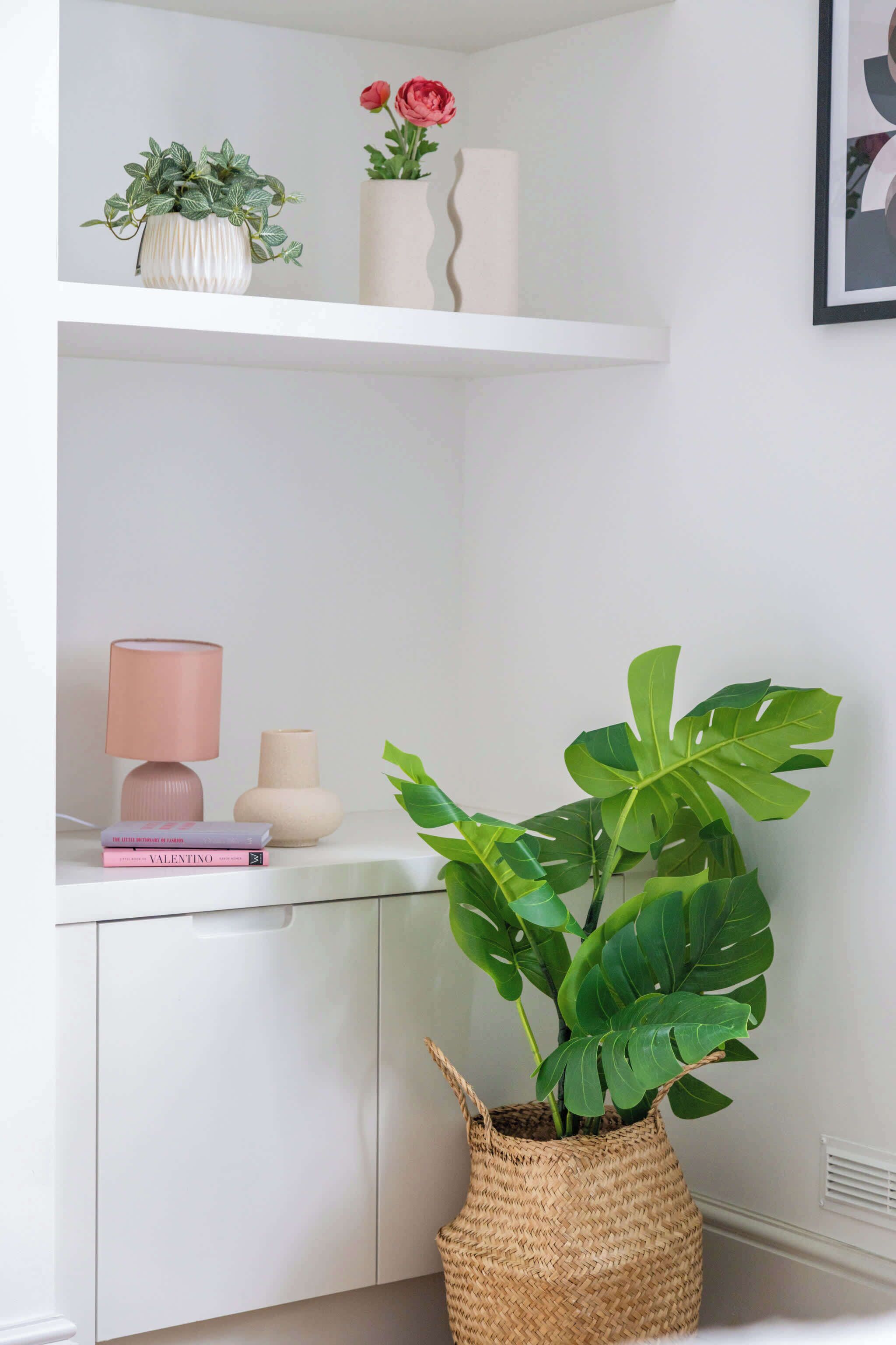 Una estant blanca amb objectes decoratius, una làmpada, un llibre i una planta verda gran en una cistella trenada.