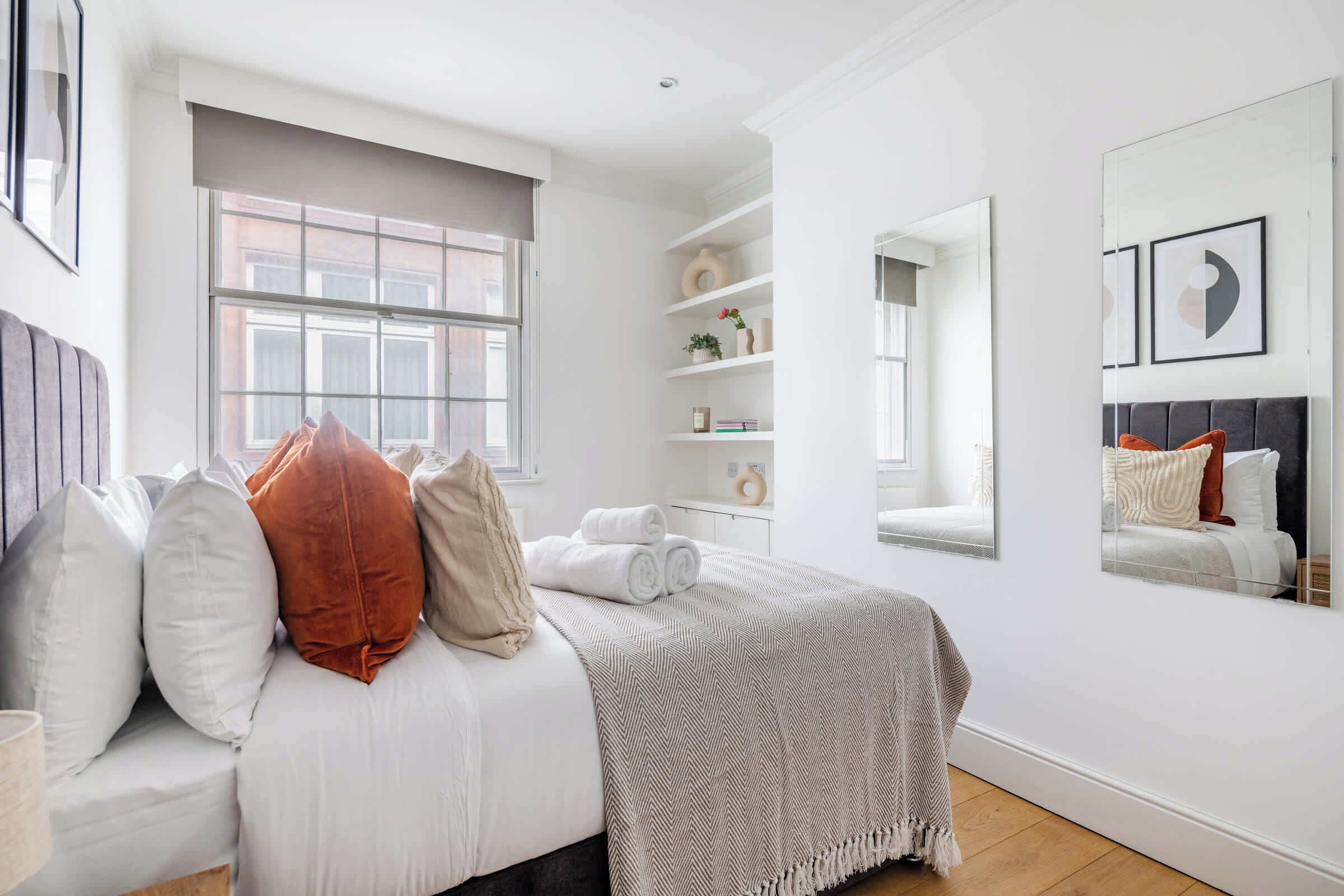 A bedroom with a bed, a window, a mirror, and a shelf with decorative items.