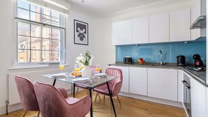 Een moderne keuken met witte kasten, een glazen eettafel, roze stoelen en een uitzicht op de stad door grote ramen.