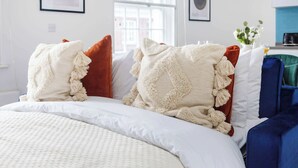 Una cama bien hecha con ropa de cama blanca y beige, una ventana con vistas a edificios y una silla azul al fondo.