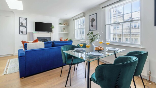 Una sala de estar moderna con un sofá azul, sillas verdes, una mesa de comedor de vidrio y un televisor en la pared.