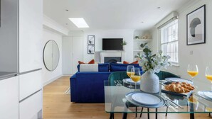 Una sala de estar moderna con un sofá azul, una mesa de comedor con platos, vasos y croissants, y un televisor de pantalla plana en la pared.