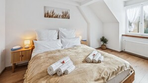 Een slaapkamer met een bed, nachtkastje, lamp, raam met gordijnen en een aan de muur hangend schilderij.