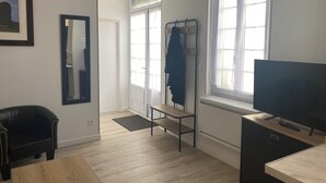 Una habitación con piso de madera, un sillón de cuero negro, un televisor sobre un mueble de madera, un perchero con una chaqueta y una puerta que da a otra habitación.