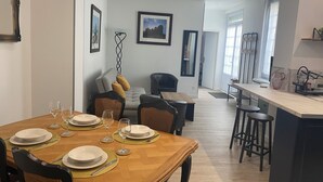 Un comedor con una mesa de madera dispuesta para cuatro personas, un sofá con cojines, una silla negra y una zona de cocina con un taburete de bar.