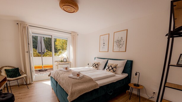 A bedroom with a bed, a chair, a small table, and a view through a glass door. - Come4Stay Passau - 2 Zimmer - bis zu 4 Gäste by Interhome (Passau)