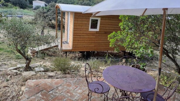 En træhytte med et lille vindue, omgivet af en terrasse med metalstole og et bord, beliggende i et naturligt landskab.