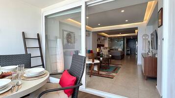 A dining area with a table set for two, a chair with a red cushion, and a sliding glass door leading to a living room with a sofa and a coffee table.