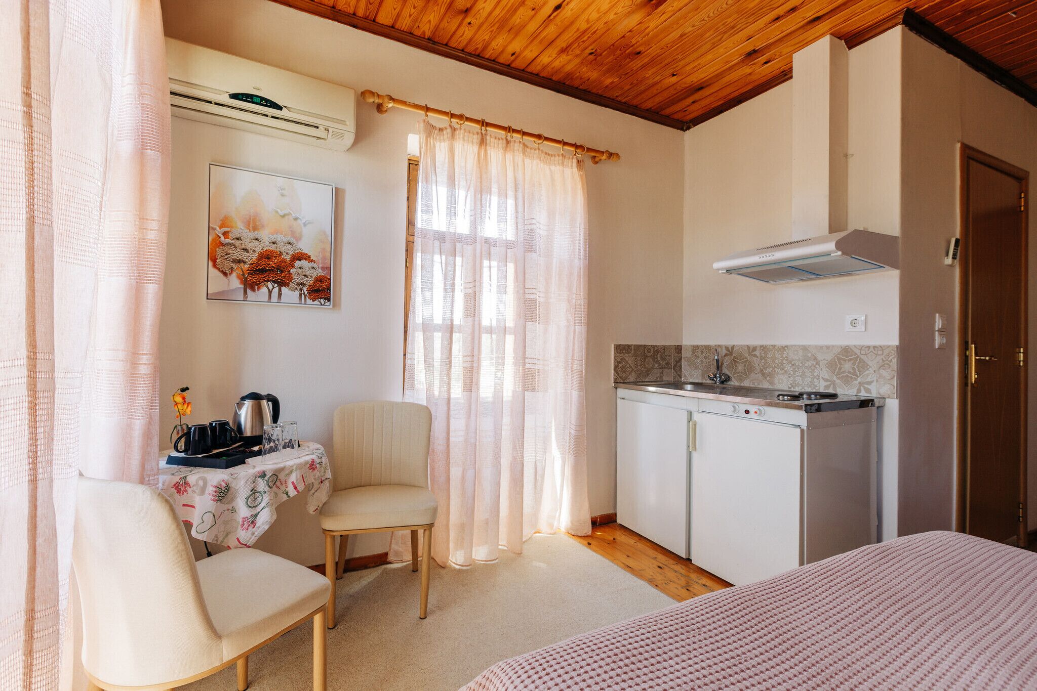 Una habitación con cama, una mesa de comedor con sillas, una kitchenette y una ventana con cortinas traslúcidas.