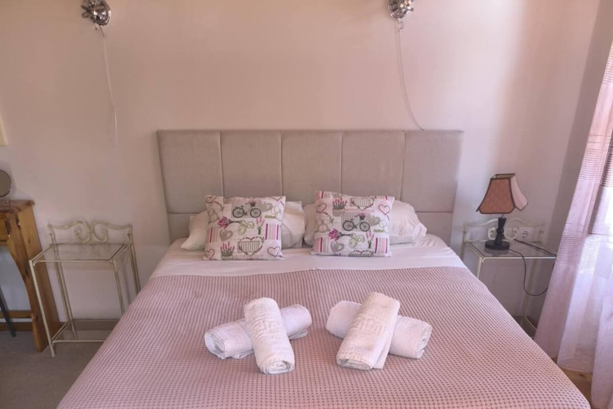 A neatly made bed with a checkered bedspread, pillows with a floral pattern, and rolled towels on the bed.