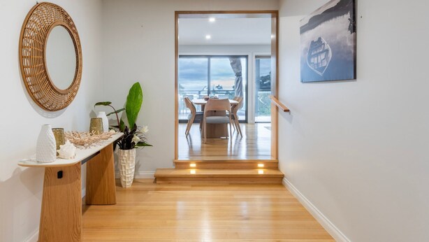 Un interior moderno con suelo de madera, un espejo redondo, una zona de comedor con mesa y sillas, y una escalera que lleva a un balcón al aire libre.