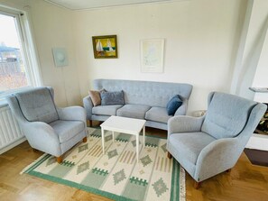 Ein Wohnzimmer mit grauem Sofa, zwei Sesseln, einem weißen Couchtisch und einem gemusterten Teppich.