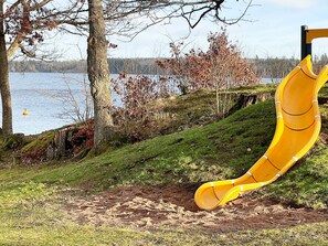 Ein Spielplatz mit Rutsche, Bäumen und einem Wasserspiegel im Hintergrund.