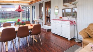 Eine überdachte Terrasse mit Holzboden, einem Esstisch mit Stühlen, einer weißen Kommode mit Spiegel und einem Fenster mit Vorhängen.