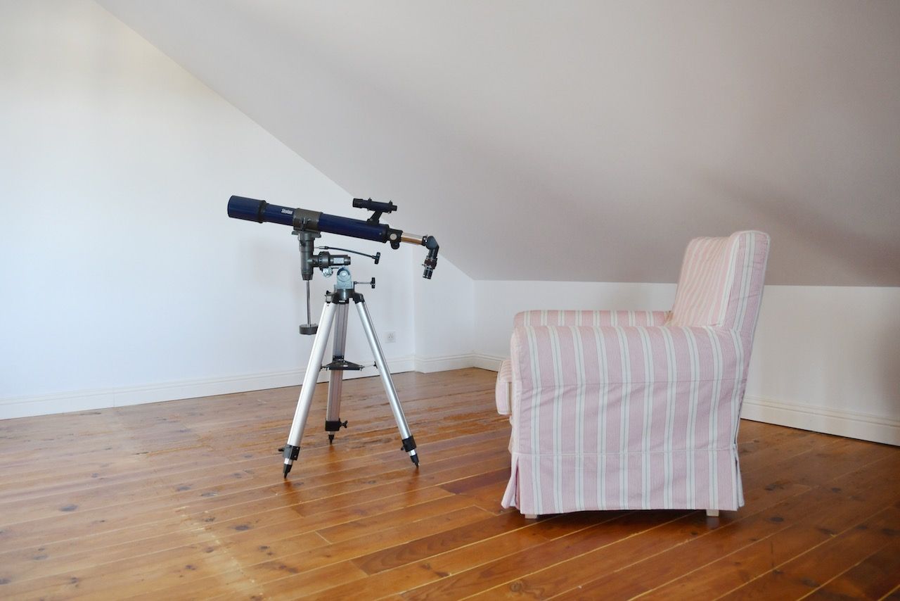 Un télescope posé sur un trépied, dans une pièce dont le sol est en bois et où se trouve une chaise à rayures.
