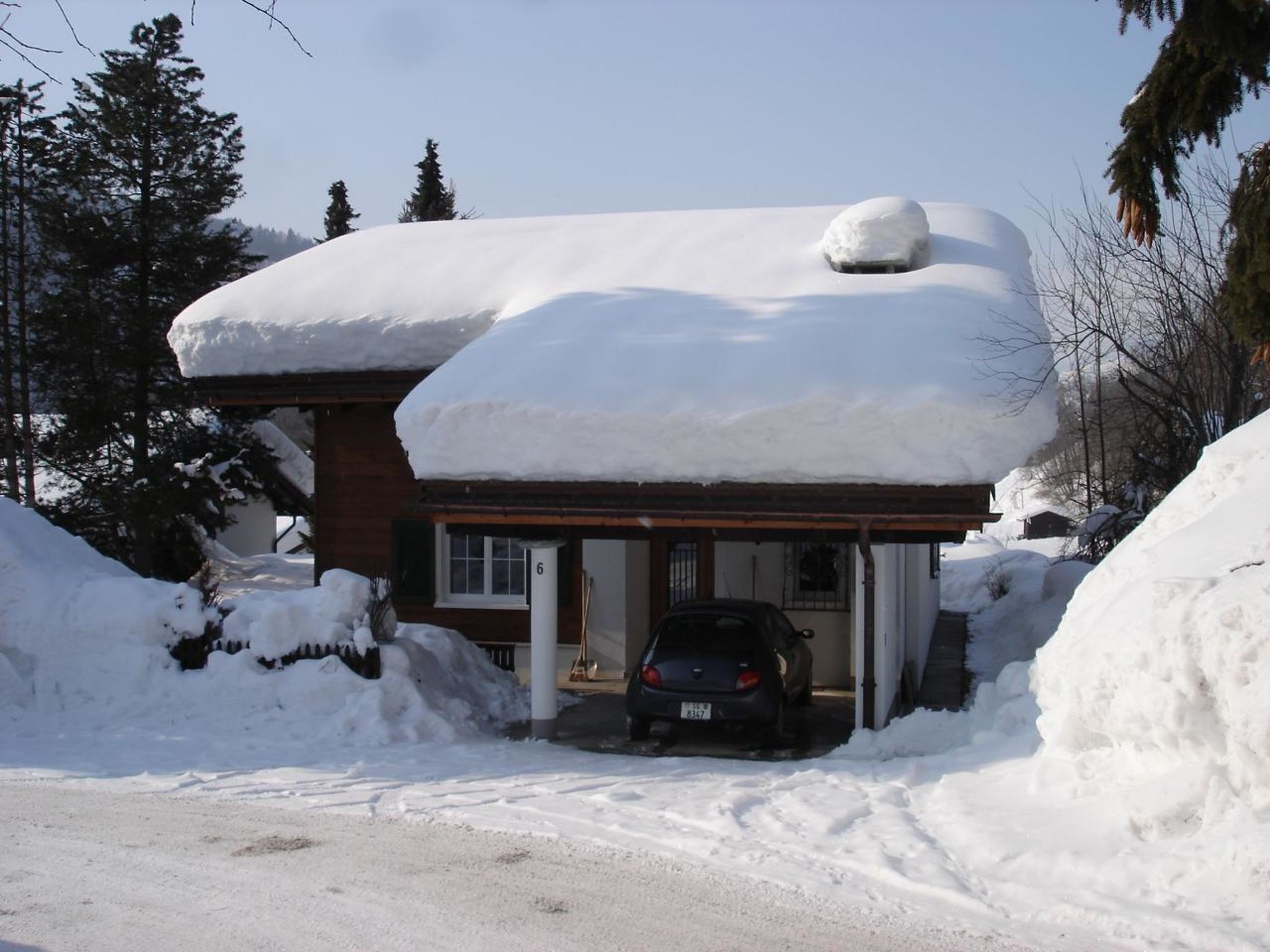 Een huis met een overdekte veranda, een auto ervoor geparkeerd, en sneeuw op het dak en de omliggende grond.