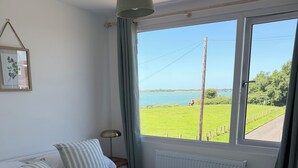 Quarto com cama, janela com vista para um campo verde e um corpo d'água, radiador e luminária de cabeceira.