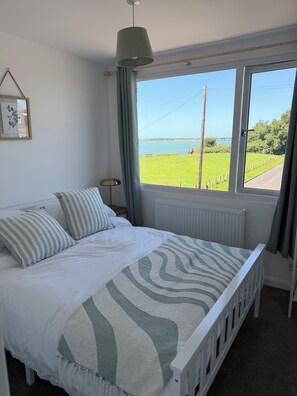 Ein Schlafzimmer mit Bett, Fensterblick auf ein grünes Feld und ein Gewässer, Heizkörper und Nachttischlampe.
