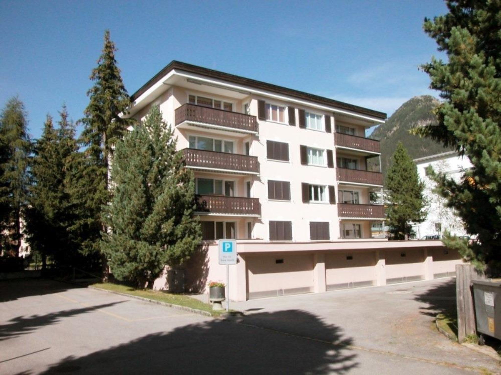 A multi-story building with balconies, a parking area, and surrounding trees.