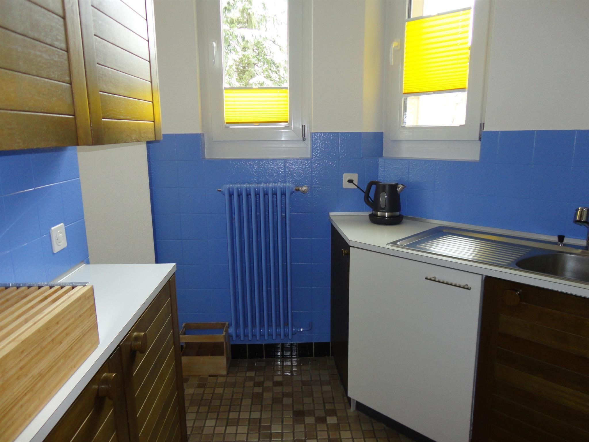 A compact kitchen with blue walls, a window with yellow blinds, a kettle, and a sink.