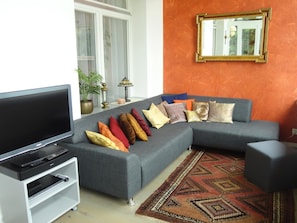 Ein Wohnzimmer mit grauem Sofa, einem Flachbildfernseher, einer weißen Unterhaltungseinheit, einem gemusterten Teppich und einem Spiegel an der Wand.