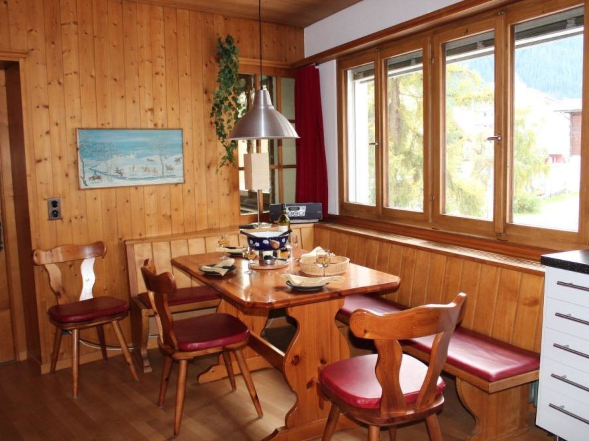 Un intérieur en bois comprenant une table à manger, des chaises, une lampe, une radio et une fenêtre donnant sur un paysage verdoyant.