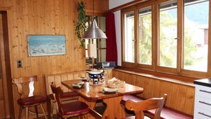 Een houten interieur met een eettafel, stoelen, een lamp, een radio en een raam met uitzicht op groen.