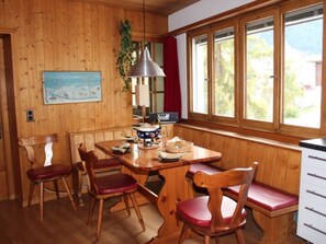 Ein Holzinnenraum mit einem Esstisch, Stühlen, einer Lampe, einem Radio und einem Fenster mit Blick auf Grünflächen.