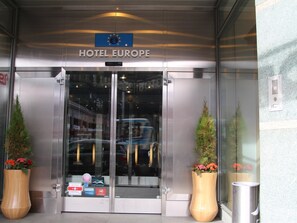 Una entrada de hotel moderna con un puerta de vidrio, un letrero que dice 'HOTEL EUROPE' y dos grandes macetas con plantas.