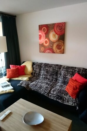 Una sala de estar con un sofá estampado en blanco y negro, cojines rojo y amarillo, una mesa de centro de madera y un cuadro abstracto colgado en la pared.