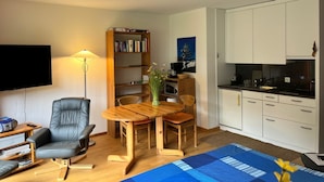 Un espacio habitable compacto con una televisión de pantalla plana, una mesa de comedor de madera con sillas, una estantería, una zona de cocina con armarios blancos y una alfombra azul.