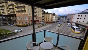 Un balcone con tavolo e sedie, affaccia su una strada con edifici e una montagna sullo sfondo.