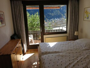 Ein Schlafzimmer mit zwei Betten, einer Holzkommode, einem Fenster mit Blick auf eine Stadt und Berge sowie einem Balkon mit Geländer.