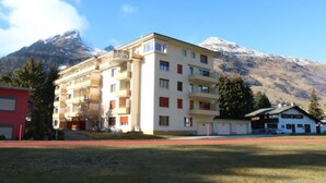 Un immeuble à plusieurs étages doté de balcons, un bâtiment rouge sur la gauche et une montagne en arrière-plan.