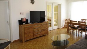 Un salon comprenant un téléviseur, un meuble en bois, une table basse en verre et un coin repas avec des chaises.