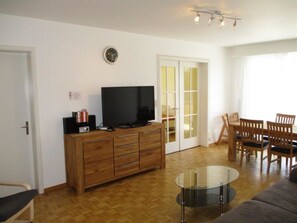 A living room with a TV, wooden cabinet, glass coffee table, and a dining area with chairs. - Bündalodge 1015 Appartement/Fewo, Dusche oder Bad, WC 4-Zimmerwohnung by Interhome (Davos)
