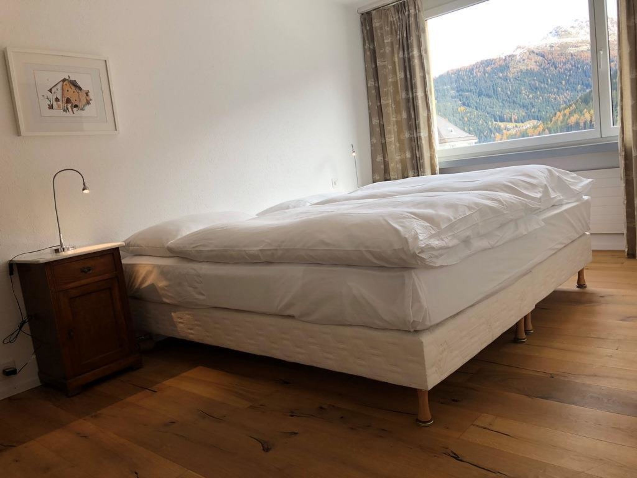Een slaapkamer met een bed, houten vloer, een nachtkastje, een lamp, een raam met uitzicht op een berg en een lijst met een afbeelding aan de muur.