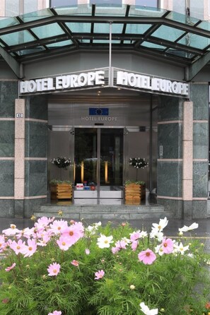 La entrada del Hotel Europe, con un toldo de vidrio, un cartel sobre las puertas y un parterre de flores al frente.