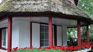 A traditional thatched-roof structure with a red and white exterior, surrounded by a well-maintained hedge and vibrant red flowers.
