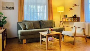 A living room with a sofa, a small table with a bowl of fruit, a chair, a bookshelf, and a lamp.