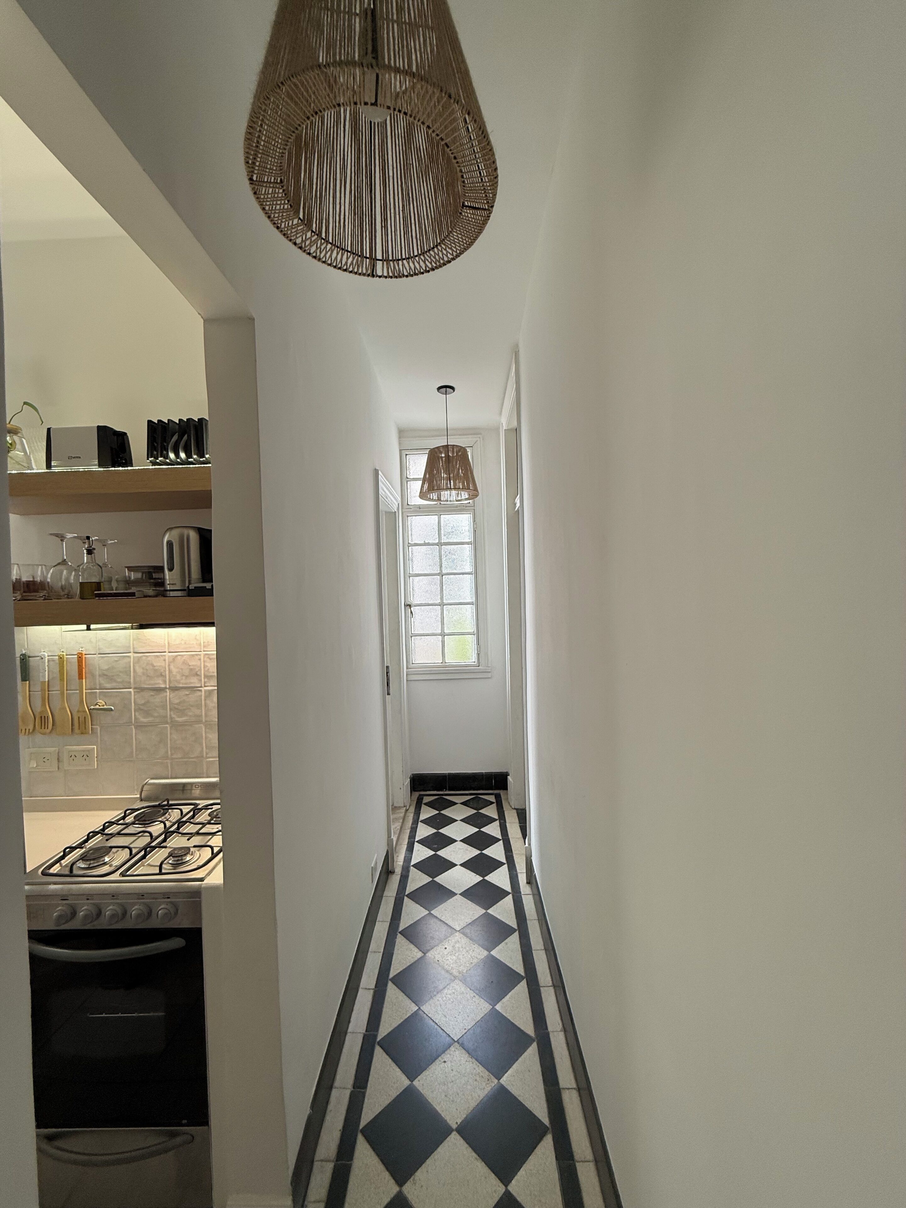 A narrow hallway with a checkered floor, a hanging light fixture, and a kitchen area with appliances. - Cozy 1-bedroom apartment in beautiful Buenos Aires with AC (Buenos Aires)