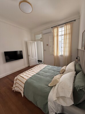 Un dormitorio con cama, televisor, ventana con cortinas y puerta.